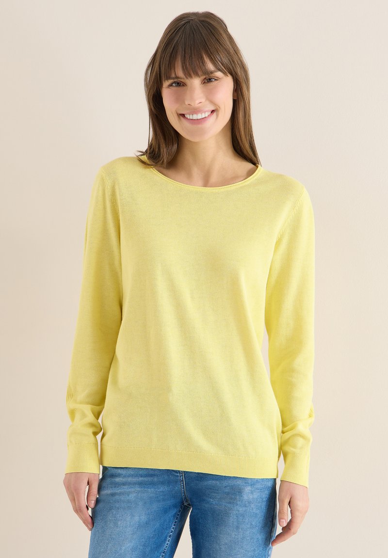 Femme aux cheveux bruns portant un pull jaune à manches longues et un jean bleu, souriante, debout devant un fond neutre.