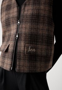 Veste zippée marron à carreaux avec deux poches devant, dont une brodée avec le mot "amour", portée par-dessus des vêtements noirs.