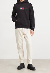 Black hoodie with front pocket, featuring "TOMMY HILFIGER" logo in red, white, and blue. Paired with cream-colored jeans and black boots.