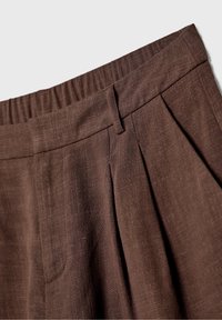 Pantalon en lin marron avec taille élastique, passants de ceinture et plis sur le devant, sur un fond clair uni.