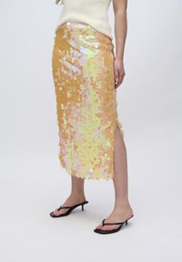 Gouden pailletten midi-rok met een glanzende textuur, voorzien van een zijsplit en cirkelpatronen, gecombineerd met zwarte hakken flip-flops.