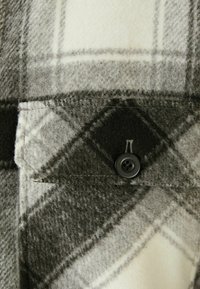 Grå och krämfärgad rutigt ulltyg med en ficka som har en svart knapp, visad med mjuk textur och en något fluffig yta.