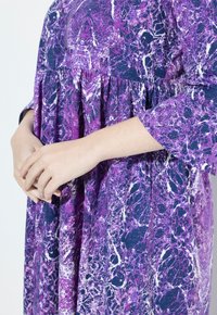 Robe à motif violet et marine avec une taille froncée, des manches trois-quarts et une coupe ample. Le tissu présente une texture marbrée.