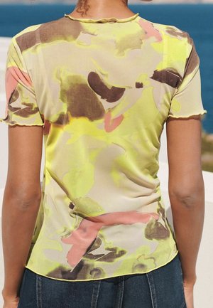 Personne portant un haut à manches courtes semi-transparent avec un motif abstrait jaune, marron et rose, debout en plein air près de l'eau.