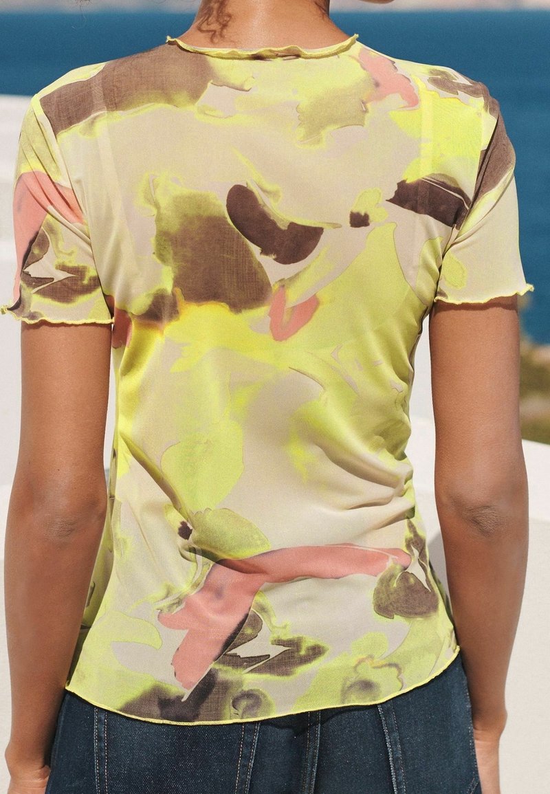 Personne portant un haut à manches courtes semi-transparent avec un motif abstrait jaune, marron et rose, debout en plein air près de l'eau.