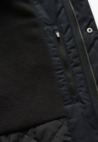 Zwarte jas met een fleece-interieur, een ritszak, gestructureerde stof en een gladde buitenkant met metallic accenten.