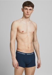 Marineblauwe katoenen boxershorts met een witte gebrandmerkte tailleband. Eenvoudig ontwerp, aansluitende vorm en zachte textuur. Model poseert tegen een neutrale achtergrond.