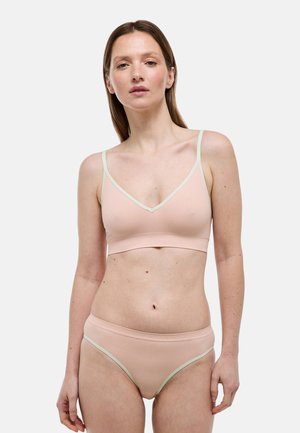 Femme aux cheveux bruns lisses portant un bralette côtelé rose clair et des sous-vêtements assortis avec un bord vert pâle, posant devant un fond blanc.
