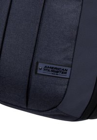 Zaino in tessuto nero testurizzato con cuciture rinforzate, caratterizzato da una forma rettangolare e una patch del logo American Tourister sul lato.
