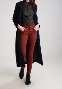 Long navy coat over a dark green button-up shirt, paired with red slim-fit pants and black lace-up boots. Smooth textures and tailored fit.