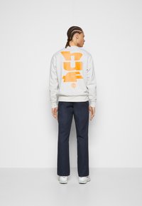 Light gray sweatshirt with large orange "HUF" logo on the back, paired with dark navy pants and white sneakers. Plain background.