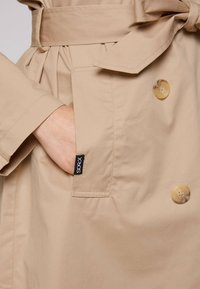 Cappotto trench beige con vita cinturata, due grandi bottoni e una tasca laterale con etichetta logo. Tessuto morbido con una leggera lucentezza.