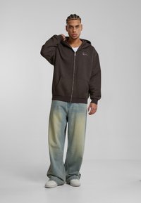 Sudadera con cremallera de color marrón, con un bolsillo tipo canguro y un logotipo bordado. El modelo lleva puestos unos vaqueros de pierna amplia en azul claro, con una textura desgastada, y zapatillas blancas.