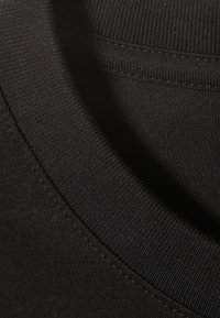 Černá bavlněná tkanina trička, detail s žebrovaným límečkem. Hladká textura s viditelným šitím podél okrajů.