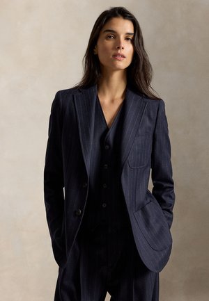 Femme aux longs cheveux foncés portant un costume à rayures sombres avec veste et gilet, debout les mains dans les poches devant un fond uni.