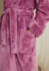 Halat de baie roz din pluș, cu centură de prins, mâneci lungi și buzunare frontale. Textură moale, cu finisaj neted și luciu subtil.
