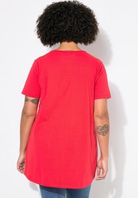 Rode katoenen T-shirt met korte mouwen, ronde hals en een iets verlengde achterkant. Kenmerkt zichtbare tatoeages op de armen.