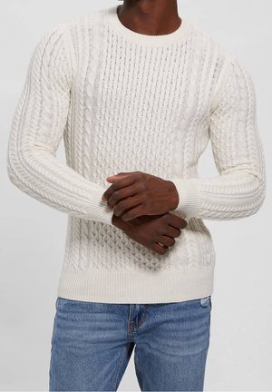 Cremefarbener Zopfmuster-Pullover mit rundem Ausschnitt, Rippbündchen und strukturierten Musterungen an den Ärmeln und dem Oberkörper. Kombiniert mit blauen Jeans.