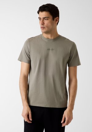 Uomo che indossa una maglietta verde oliva a maniche corte con il logo "GUESS" e pantaloni neri, guarda verso sinistra su uno sfondo semplice.