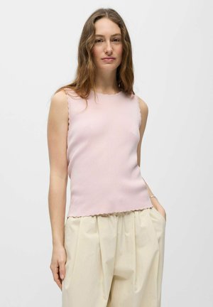 Femme aux longs cheveux ondulés, portant un haut sans manches côtelé rose clair et un pantalon large crème, debout avec une main dans la poche.