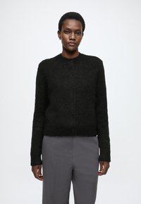 Czarny sweter z teksturą, o okrągłym dekolcie, z przodu zapinany na guziki, z długimi rękawami, zestawiony z szarymi spodniami. Minimalistyczny design, bez wzorów.