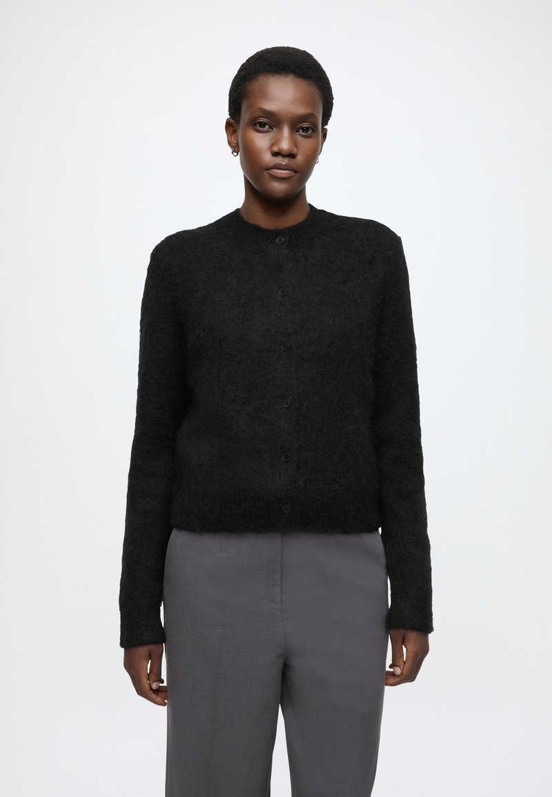 Czarny sweter z teksturą, o okrągłym dekolcie, z przodu zapinany na guziki, z długimi rękawami, zestawiony z szarymi spodniami. Minimalistyczny design, bez wzorów.