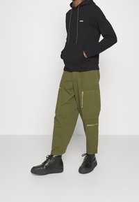 Sweat à capuche noir avec logo blanc et cordon de serrage, associé à un pantalon cargo vert olive avec poches zippées et des baskets noires.