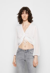 Top curto branco com decote em V, detalhe franzido na frente e mangas longas e transparentes. Combinado com jeans cinza claro. Tecido suave com um leve brilho.