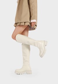 Stivali bianchi al ginocchio con finitura lucida, suola spessa e design elegante, abbinati a un blazer beige e una gonna plissettata.