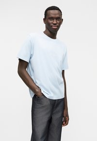 Helesinine puuvillane lühikeste varrukatega t-särk ümmarguse kaelusega ja peene tekstiga rinnal, kombineeritud tumeda halli teksapükstega.