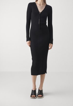 Femme portant une longue robe noire en maille côtelée avec manches longues, une fermeture éclair devant et des sandales noires ouvertes à enfiler.