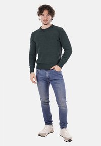 Maglione lavorato a maglia verde scuro con scollo a girocollo, abbinato a jeans azzurri e sneaker bianche. Il modello è in piedi contro uno sfondo neutro.