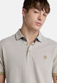 Ljusgrå polo-tröja med en texturerad tyg, tre-knapp placket och kontrasterande marinblå krage. Har en liten, broderad logotyp på bröstet.