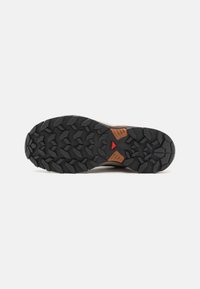 Semelle d'une chaussure de randonnée avec des crampons noirs profonds, des accents bruns et un petit triangle rouge au centre, conçue pour l'adhérence et la durabilité.