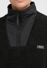 Black fleece pullover with a high collar, featuring a zippered front. Smooth panel at the collar, textured body, and logo badge on the side.