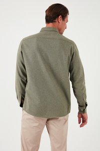 Camisa verde de tejido con cuello y mangas largas, vista desde la parte de atrás. Pantalones beige claros visibles debajo de la camisa.