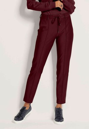 Slim-Fit burgunderrote Hose mit Kordelzug in der Taille, getragen mit schwarzen Low-Top-Sneakers an einer stehenden Person.