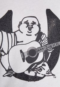 Biała koszulka z czarną grafiką uśmiechniętej postaci grającej na gitarze akustycznej, ozdobiona czarnymi kamieniami strassowymi w projekcie.