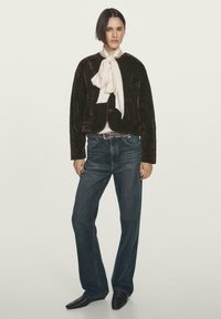 Brun fløjlsjakke med rund hals, hvidt tørklæde og mørkeblå jeans. Bootcut-design med et skinnende nittebælte og sorte sko.