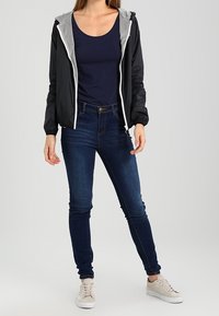 Černá lehká bunda se šedou kapucí, námořnicky modrý top na ramínka, modré slim džíny a béžové tenisky. Zaměření na ležérní, vrstvený outfit.