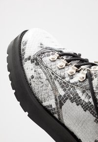 Botas con estampado de serpiente blanco y negro, con una superficie texturizada, cordones negros, ojales metálicos y una gruesa suela de goma negra.