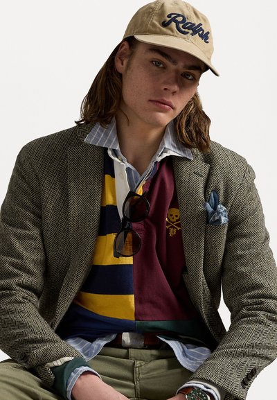 Fiskebensblazer i grønn og grå, lagd over en stripete skjorte med knapper og en fargeblokkert polo. Beigefarget caps med "Ralph"-logo. Solbriller hvilende på skjorten.