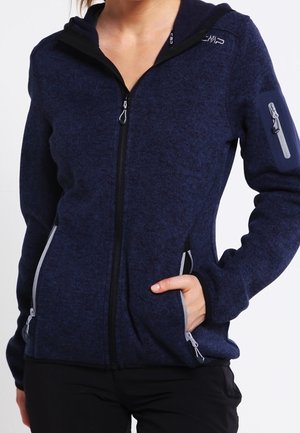 Mujer que lleva una chaqueta de forro polar azul marino con cremallera, con bolsillos con cremallera y un pequeño bolsillo con cremallera en la parte superior de la manga izquierda.