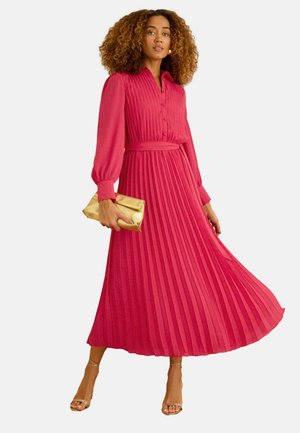 Göndör hajú nő, aki hosszú ujjú, pliszírozott piros ruhát visel, arany borítéktáskát tart, és pántos arany magas sarkú cipőt visel, lépés közben pózol.