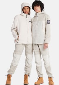 Beige Fleece-Jacke mit Kapuze und Reißverschluss, kombiniert mit passenden hellen Hosen mit Paneelakzenten. Braune Stiefel vervollständigen das Outfit.