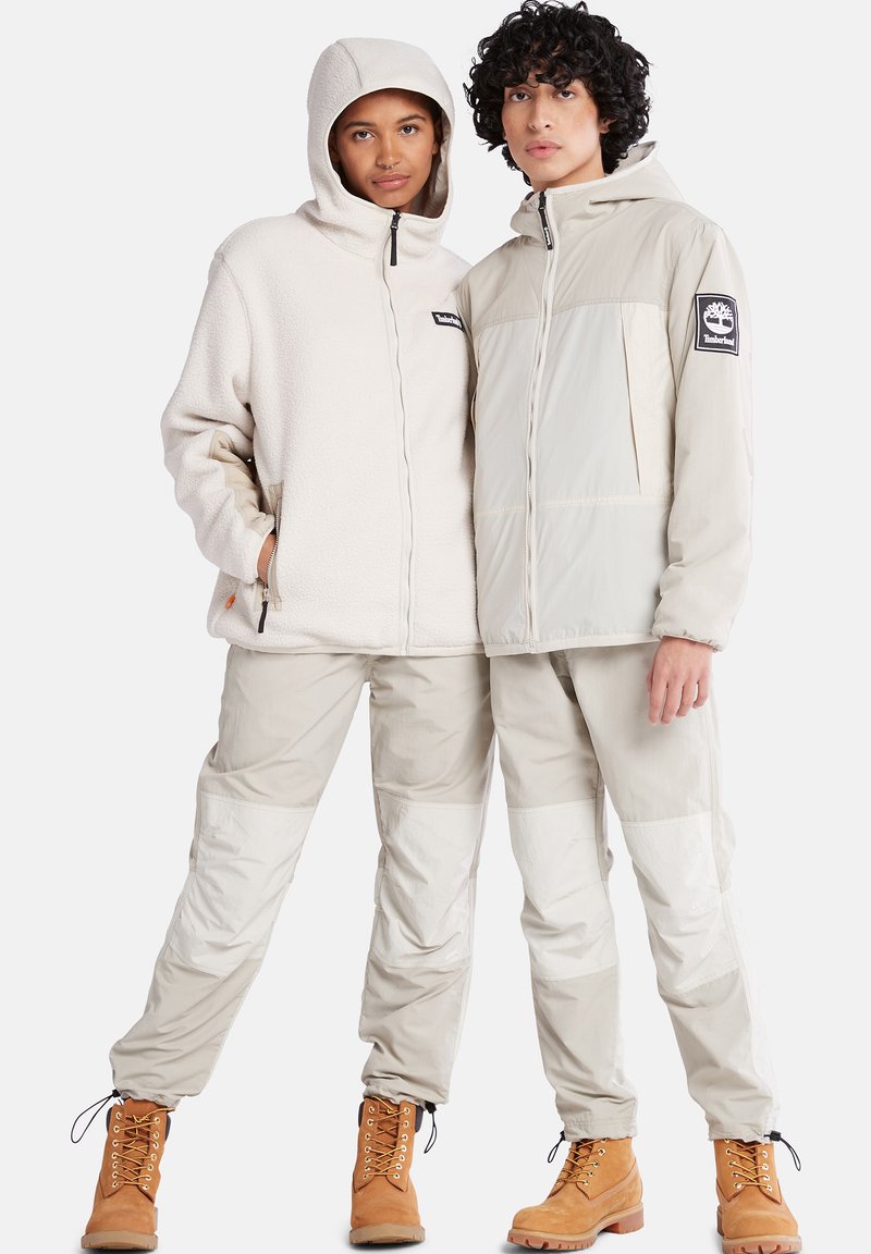 Beige Fleece-Jacke mit Kapuze und Reißverschluss, kombiniert mit passenden hellen Hosen mit Paneelakzenten. Braune Stiefel vervollständigen das Outfit.