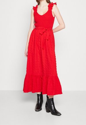 Woman wearing a sleeveless red midi dress with ruffled straps and a waist tie, paired with black textured ankle boots.