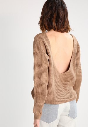 Femme aux cheveux foncés mi-longs portant un pull en maille marron ample avec un dos profondément ouvert et un pantalon clair avec des poches grises.