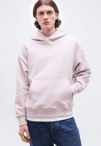 Jeune homme aux cheveux bruns mi-longs portant un sweat à capuche rose clair et un jean bleu, se tenant devant un fond blanc uni.