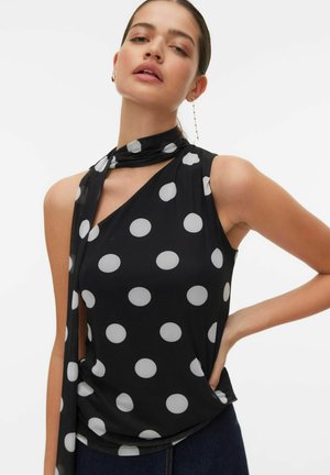 Vrouw draagt een zwart mouwloos topje met grote witte stippen en een bijpassende sjaal om de nek geknoopt, poseert met één hand op de heup.